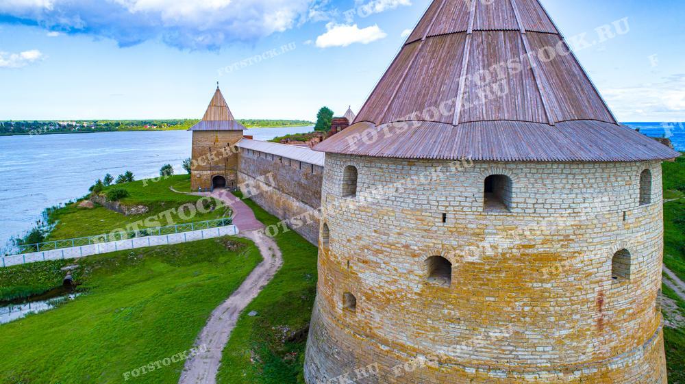 На фотографии представлены оборонительные сооружения крепости Орешек, расположенные на острове на реке Неве. Изображены характерные башни крепости с конусообразными крышами и каменные стены, окружающие её. Фотография сделана с высоты, позволяя оценить уникальное сочетание исторической архитектуры и природного ландшафта. Это изображение может быть использовано в туристических буклетах, исторических обзорах, образовательных материалах и как элемент визуального оформления статей о культурном наследии.